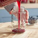 A hand pouring red liquid into a 26 oz. square glass mason jar.