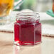 A 1.5 oz. hex glass jar of red liquid sitting on a table next to a glass of juice.