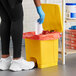 A person wearing blue rubber gloves putting white paper into a yellow Lavex rectangular step-on trash can.