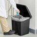 A person in a lab coat throwing a blue bag into a Lavex black rectangular step-on trash can.