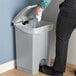 A person using their foot to open a Lavex gray rectangular step-on trash can to put a white plastic bag inside.