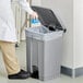 A person in a white coat and khaki pants putting a blue plastic bag into a gray Lavex step-on trash can.