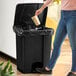 A woman in jeans using a Lavex black rectangular step-on trash can to throw away a paper cup.