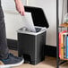 A person standing next to a Lavex black rectangular step-on trash can with a white paper in hand opening the lid to throw it away.