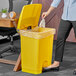 A woman's hand throwing a bag into a yellow Lavex step-on trash can.