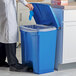 A man in a white lab coat using a gloved hand to open a blue Lavex step-on trash can on a counter with a plastic bag inside.