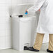 A man in a white lab coat using blue gloves to step on a white Lavex rectangular step-on trash can.