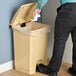 A man standing next to a Lavex tan rectangular step-on trash can with a plastic bag in it.