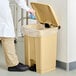 A person in a lab coat and gloves putting a blue plastic bag inside a tan Lavex rectangular step-on trash can.