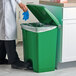 A person in a white lab coat putting a plastic bag in a green Lavex step-on trash can.