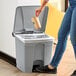 A hand holding a coffee cup pours coffee into a Lavex rectangular step-on trash can in a corporate office cafeteria.