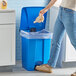 A woman's legs in jeans and shoes throwing a paper bag into a blue Lavex step-on trash can.