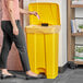 A woman standing next to a Lavex yellow rectangular step-on trash can.