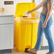 A woman standing in a kitchen and putting a plastic bag in a yellow Lavex step-on trash can.