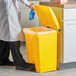 A person in a white lab coat and gloves putting a blue glove into a yellow Lavex step-on trash can.