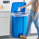 A woman using a Lavex blue rectangular step-on trash can to throw tissue in a plastic bag inside.