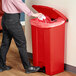 A person in black pants uses a white cloth to put a bag in a red Lavex step-on trash can.