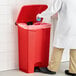 A person in a white lab coat putting a blue glove into a red Lavex step-on trash can.