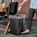 A woman using a Lavex black rectangular step-on trash can to throw away paper in a corporate office.