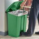 A person standing next to a green Lavex step-on trash can with a plastic bag puts a cup inside.