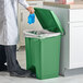 A person in a white lab coat putting a blue glove into a green Lavex step-on trash can.