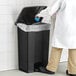 A person in a lab coat throws a blue glove into a Lavex rectangular step-on trash can.