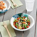 A bowl of salad with croutons and tomatoes in a Choice heavy weight paper bowl on a table with a wooden fork and clear plastic cup of water.