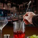 A hand using a Barfly stainless steel shark end bar spoon to stir a cocktail in a glass of red liquid.