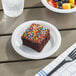 A Choice white plastic plate with chocolate cake with sprinkles and a bowl of fruit on it next to a fork.