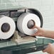 A hand pushing a Lavex jumbo roll of toilet paper on a toilet paper holder.