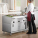 A woman in a red apron and gloves using a ServIt electric steam table in a commercial kitchen.