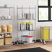 A Regency chrome wire shelving unit in a school kitchen with metal containers and buckets on the shelves.