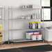 A Regency metal wire shelving unit in a professional kitchen with white containers on the shelves and a white and red container next to it.