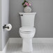 An American Standard Edgemere matte black toilet paper holder on a white bathroom wall above a toilet with a vase of flowers on top.