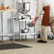 A person wearing an apron and standing next to a Regency stainless steel mixer table holding a metal bowl.