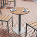 A Flash Furniture natural faux teak and metal table with plates and wine glasses on it on a brick patio.