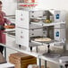 A man in an apron using a Cooking Performance Group countertop conveyor oven to cook a pizza.