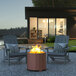 A Flash Furniture Titus bronze steel portable fire pit on a table with two chairs and a black and white pillow in front of a house.