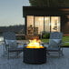 A Flash Furniture black steel fire pit with a fire in it on an outdoor patio table with chairs around it.