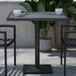 A Flash Furniture square gray wash faux teak table and two black chairs on a patio with a potted plant.