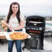 A woman holding a pizza in front of a Blackstone outdoor pizza oven on a stand.