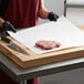 A person wearing a black glove is cutting Choice 18" freezer paper on a cutting board.