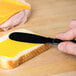 A person using a Sabert black plastic spreader to spread yellow spread on a piece of bread.