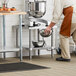 A man in a brown apron using a Regency stainless steel mixer table with a metal bowl and whisk.