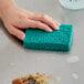 A hand holding a blue Simple Green heavy-duty scrubbing pad on a stainless steel surface.