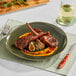 An Acopa Highlands juniper green china plate with lamb chops, vegetables, and a glass of wine on a table.