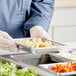 A person in a blue uniform holding a Vollrath stainless steel steam table pan full of food on a counter.
