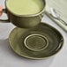 A hand holding a cup of green tea over a juniper green Acopa Highlands saucer on a table.