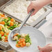 A hand using a Thunder Group perforated basting spoon to serve vegetables onto a plate.