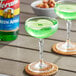 A glass of green liquid on a table next to a bottle of Torani Green Apple syrup.